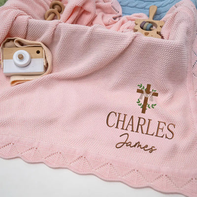 Pink knitted blanket with embroidered name and cross design, surrounded by baby toys.