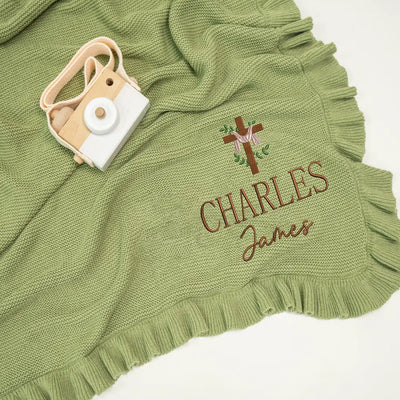 Green knitted blanket with embroidered name and cross design, accompanied by a small camera toy.