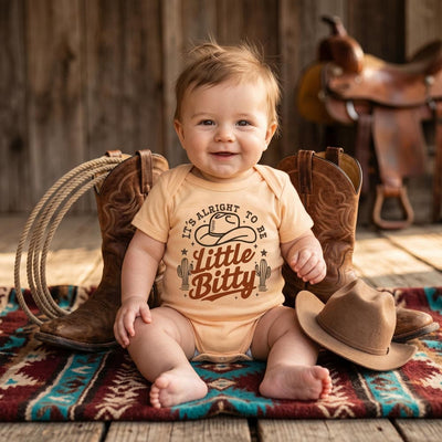 Baby wearing a cowboy-themed onesie sitting on a rug with cowboy boots and a hat in the background.