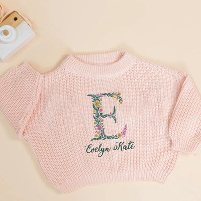 Pink knitted sweater with floral letter 'E' and text on a beige background