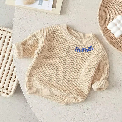 Beige baby sweater with 'Thomas' embroidered on it, placed on a light surface.