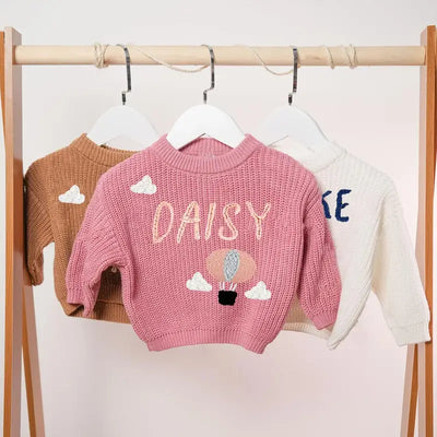 Pink children's sweater with 'DAISY' text and cloud design on a rack.