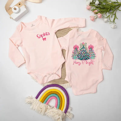 Two pink baby onesies with Christmas designs on a light gray surface with decorative items.