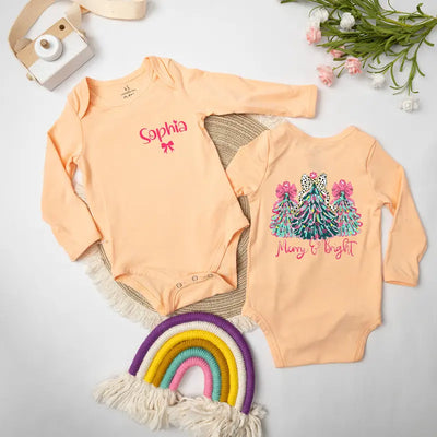 Two peach-colored baby onesies with Christmas-themed designs on a light gray surface.