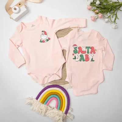 Two pink baby onesies with Christmas-themed designs on a light gray surface with decorative items.