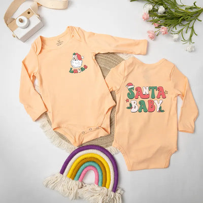 Two peach-colored baby onesies with Christmas-themed designs on a light gray surface.