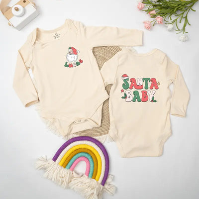 Two baby onesies with Christmas designs on a white surface with a rainbow toy and flowers.