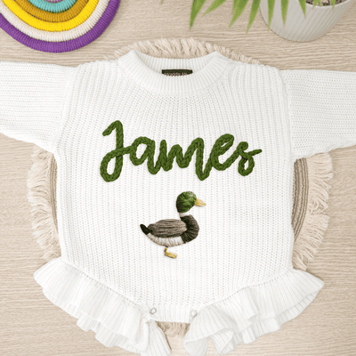 White baby romper with 'James' and a duck design on a wooden surface.
