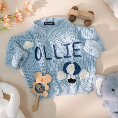 Blue knitted sweater with 'OLLIE' embroidered on it, surrounded by toys and flowers.