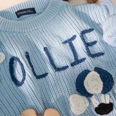 Light blue knitted sweater with 'OLLIE' embroidered on it, featuring flower designs.