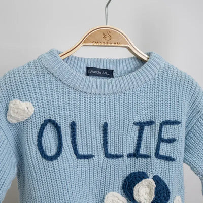 Blue sweater with 'OLLIE' embroidered on it, hanging on a wooden hanger against a gray background.