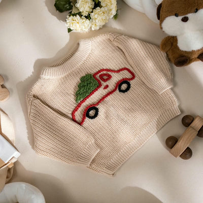 Beige knitted sweater with a red car design on a light surface with toys and flowers.