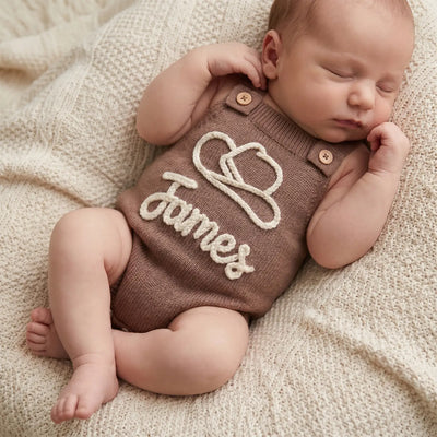 Newborn baby wearing a brown knitted romper with 'James' embroidered on it, lying on a soft beige blanket.