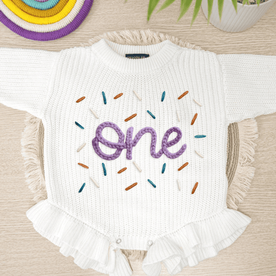 White baby romper with 'one' embroidered in purple and colorful sprinkles on a wooden surface.
