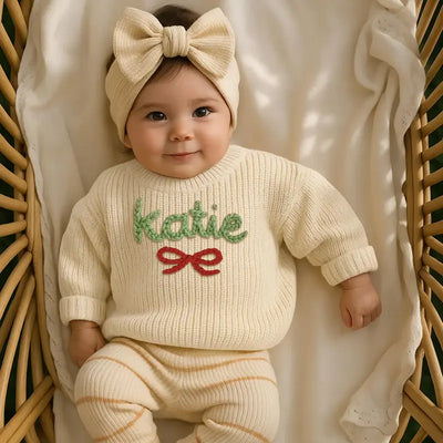 Baby wearing a cream sweater with 'katie' and a bow, sitting in a wicker basket.