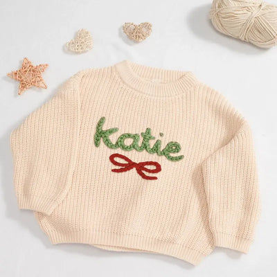 Beige sweater with 'katie' embroidered in green and a red bow on a light gray background.