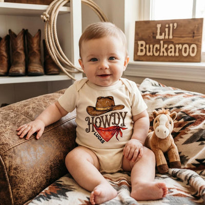 Baby wearing a 'Howdy' onesie with cowboy hat graphics, sitting on a patterned blanket with cowboy boots and a wooden sign in the background.
