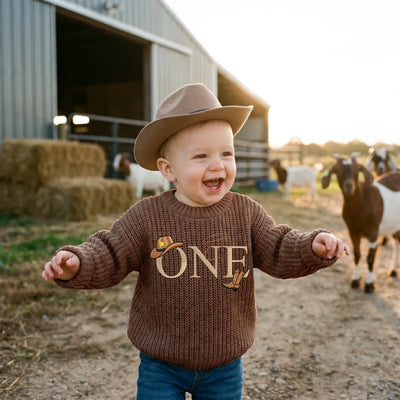 Child wearing a brown sweater with 'ONE' on it, standing in a barnyard with animals.