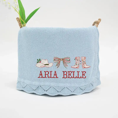 Light blue knitted blanket with embroidered designs and 'Aria Belle' text, displayed on a white background.