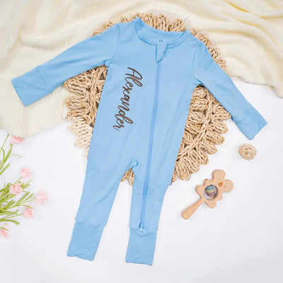 Light blue baby onesie with name 'Alexander' on a textured surface with flowers and toys.