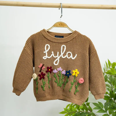 Brown sweater with embroidered flowers and 'Lyla' text on a hanger against a light background.