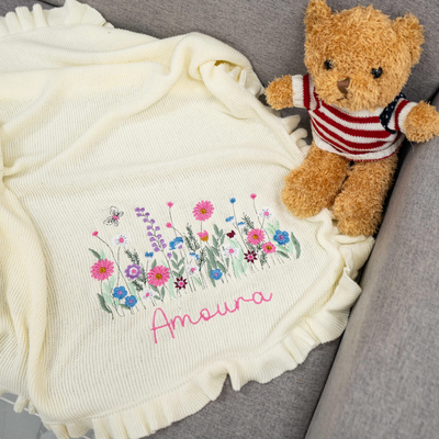 Blanket with floral design and embroidered name, accompanied by a teddy bear on a gray surface.