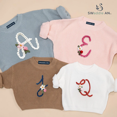 Four sweaters with embroidered letters 'A', 'E', 'J', and 'Q' on a beige background, branded as Swaddle AN.
