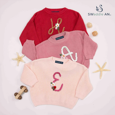 Three children's sweaters in red, pink, and light pink with decorative letters on a light background.