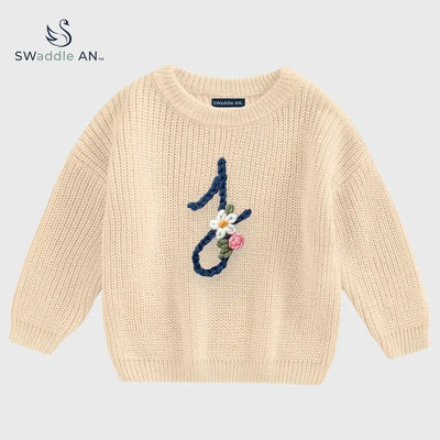 Beige sweater with floral design and number '1' on a light gray background, branded 'Swaddle AN'.