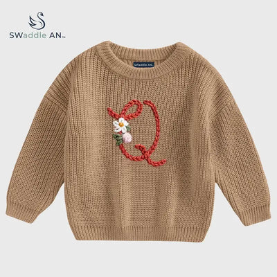 Brown knitted sweater with a decorative letter 'Q' on a light gray background, featuring the brand 'Swaddle AN'.