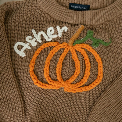 Brown sweater with embroidered orange pumpkins and white text on a plain background