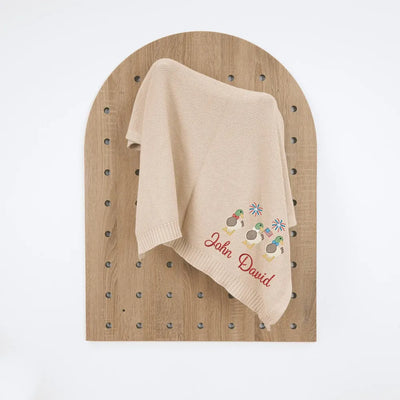 Beige baby blanket with embroidered patriotic mallard duck trio design and text on a wooden pegboard.