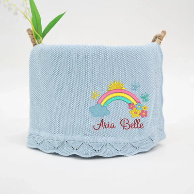 Light blue knitted blanket with embroidered rainbow and flowers, featuring the name 'Aria Belle'.