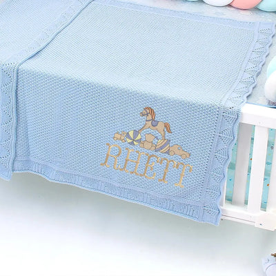 Light blue personalized baby blanket with embroidered rocking horse design and name 'Rhett' on a crib.