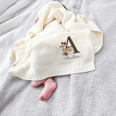 Baby wrapped in a cream personalized baby blanket with a personalized reindeer alphabet design on a gray surface