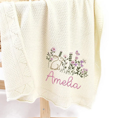 Cream personalized baby blanket with embroidered bunny floral design and name 'Amelia' on a wooden stand.