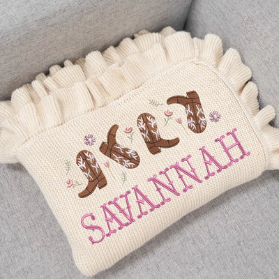 Cream personalized baby blanket with embroidered cowgirl boots and the name 'Savannah' on a gray surface.