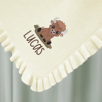 Cream personalized baby blanket with a cartoon highland cow and the name 'Lucas' on a light gray background