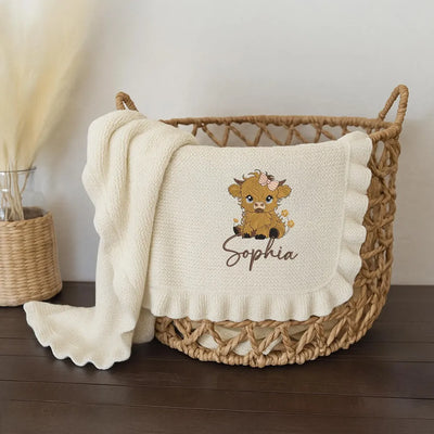 Cream personalized baby girl blanket with a cartoon highland cow design and name 'Sophia' in a wicker basket on a wooden floor.