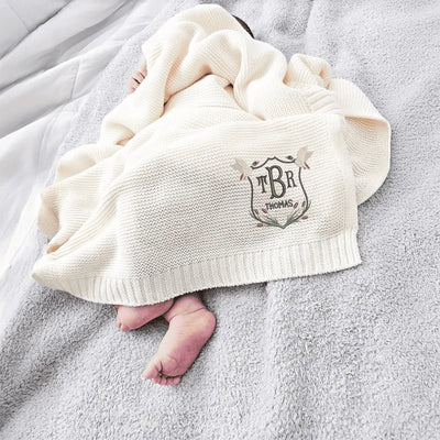 Baby wrapped in a cream personalized blanket with a mallard duck crest logo and text, lying on a gray surface.