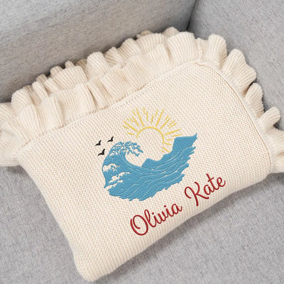 Cream personalized baby blanket with embroidered ocean waves design and text on a gray surface