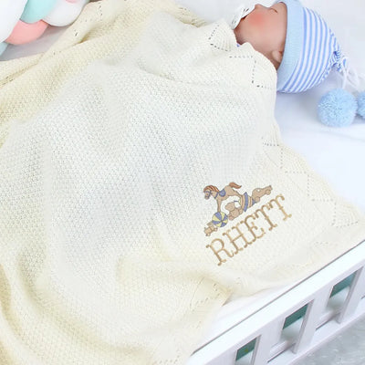 Cream personalized baby blanket with embroidered name and rocking horse design, featuring a baby in the background.
