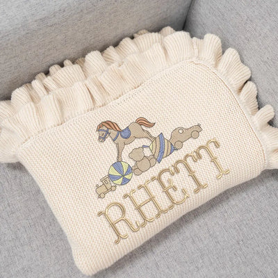Cream personalized baby blanket with embroidered rocking horse and rider design and 'Rhett' text on a gray surface.