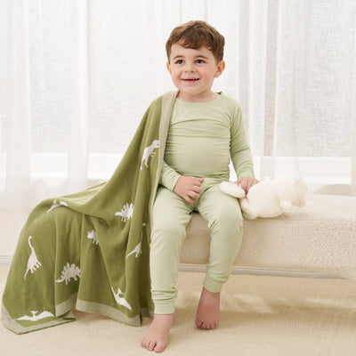 Child in green pajamas sitting on a couch with a dinosaur-patterned blanket