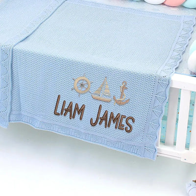 Blue personalized baby blanket with embroidered name and anchor and sailboat symbols on a crib.