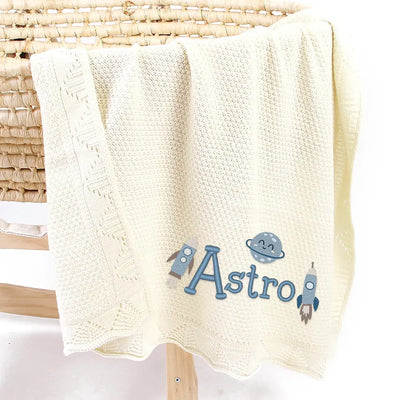 Cream personalized baby blanket with 'Astro' and astronaut designs on a wicker chair.