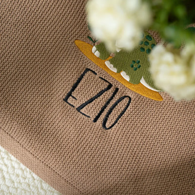 Close-up of a mocha personalized baby blanket with 'ELIO' embroidered on it, featuring a cute dinosaur design.