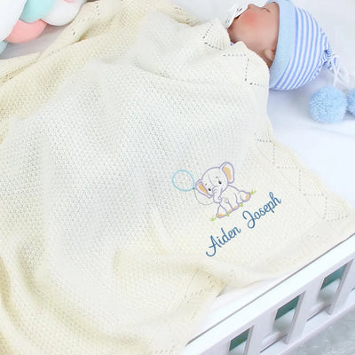 Baby in a cream personalized blanket with an embroidered elephant design and name, lying on a white surface.