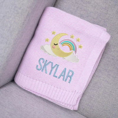 Personalized baby blanket with a rainbow, moon, and stars design and the name 'Skylar' on a gray surface.
