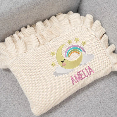 Cream personalized baby blankets with embroidered design of a moon, rainbow, and stars, and the name 'Amelia' on a gray surface.